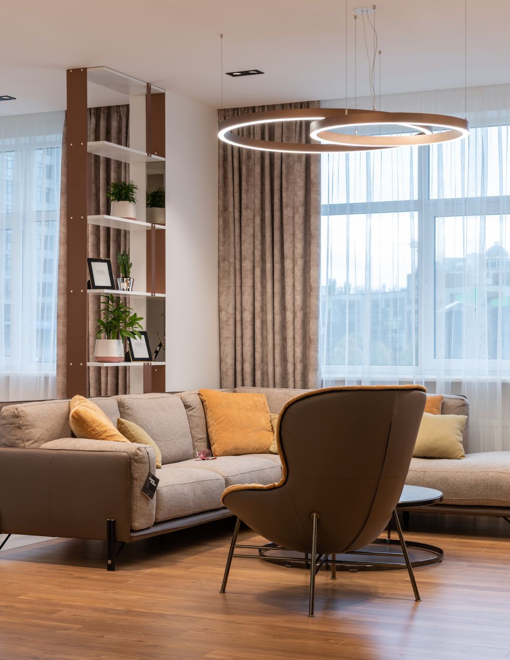 A furnished family living room in late-morning window light, fresh vacuum lanes still visible across pale wool carpet, plumped cushions on the sofa and a throw folded over the arm